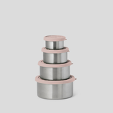 Four nested stainless steel lunch containers with dusty pink lids and rims, arranged in a stack against a solid light gray background.