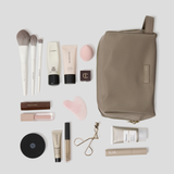 Oyster makeup travel pouch arranged on a light gray background with various makeup items including brushes, tubes, a compact, and an eyelash clip.
