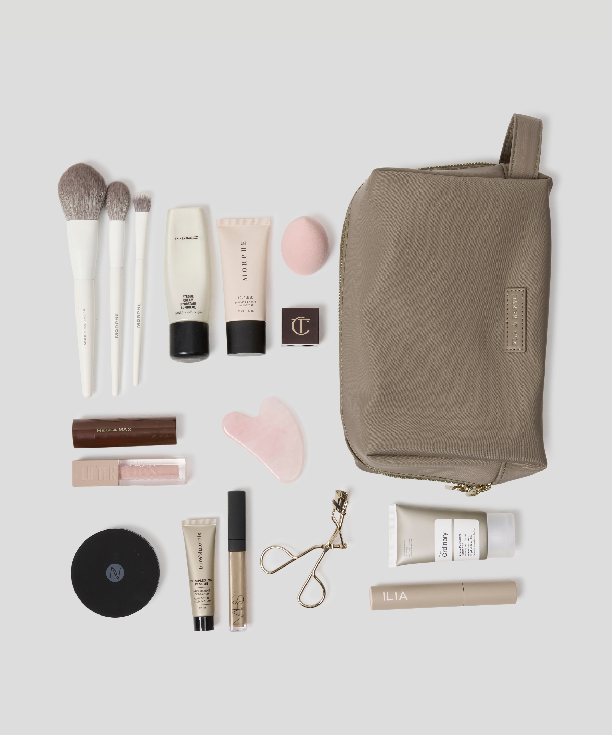 Oyster makeup travel pouch arranged on a light gray background with various makeup items including brushes, tubes, a compact, and an eyelash clip.