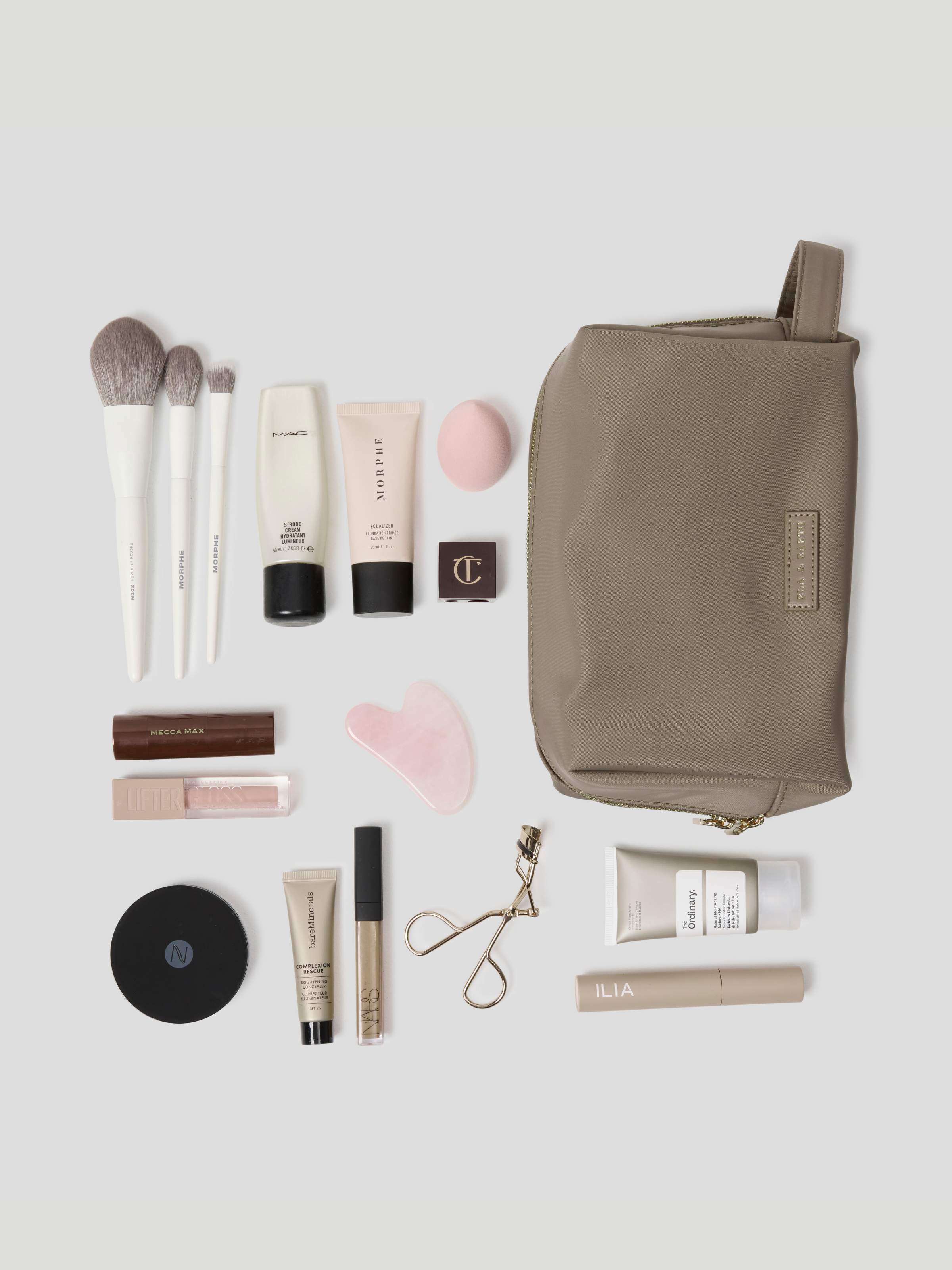 Oyster makeup travel pouch arranged on a light gray background with various makeup items including brushes, tubes, a compact, and an eyelash clip.