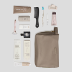 A flat-lay arrangement of toiletry and grooming items on a light gray background, including a taupe zip pouch, a black comb, a pink-handled razor, several white and beige tubes, and a small black bottle. The pouch is the largest item and appears to be the main product, with the other items styled as included accessories or companion products.