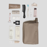 A flat-lay arrangement of toiletry and grooming items on a light gray background, including a taupe zip pouch, a black comb, a pink-handled razor, several white and beige tubes, and a small black bottle. The pouch is the largest item and appears to be the main product, with the other items styled as included accessories or companion products.