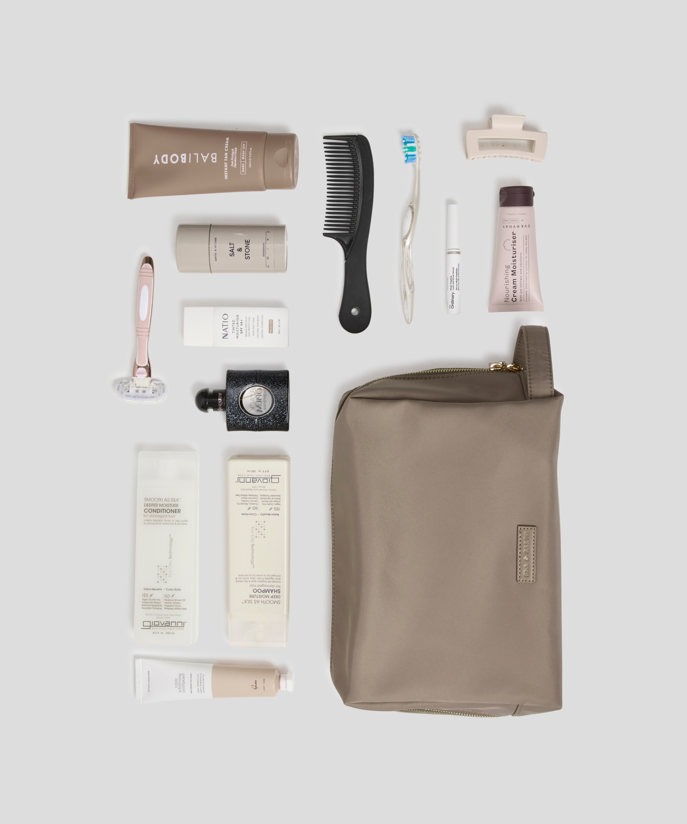 A flat-lay arrangement of toiletry and grooming items on a light gray background, including a taupe zip pouch, a black comb, a pink-handled razor, several white and beige tubes, and a small black bottle. The pouch is the largest item and appears to be the main product, with the other items styled as included accessories or companion products.