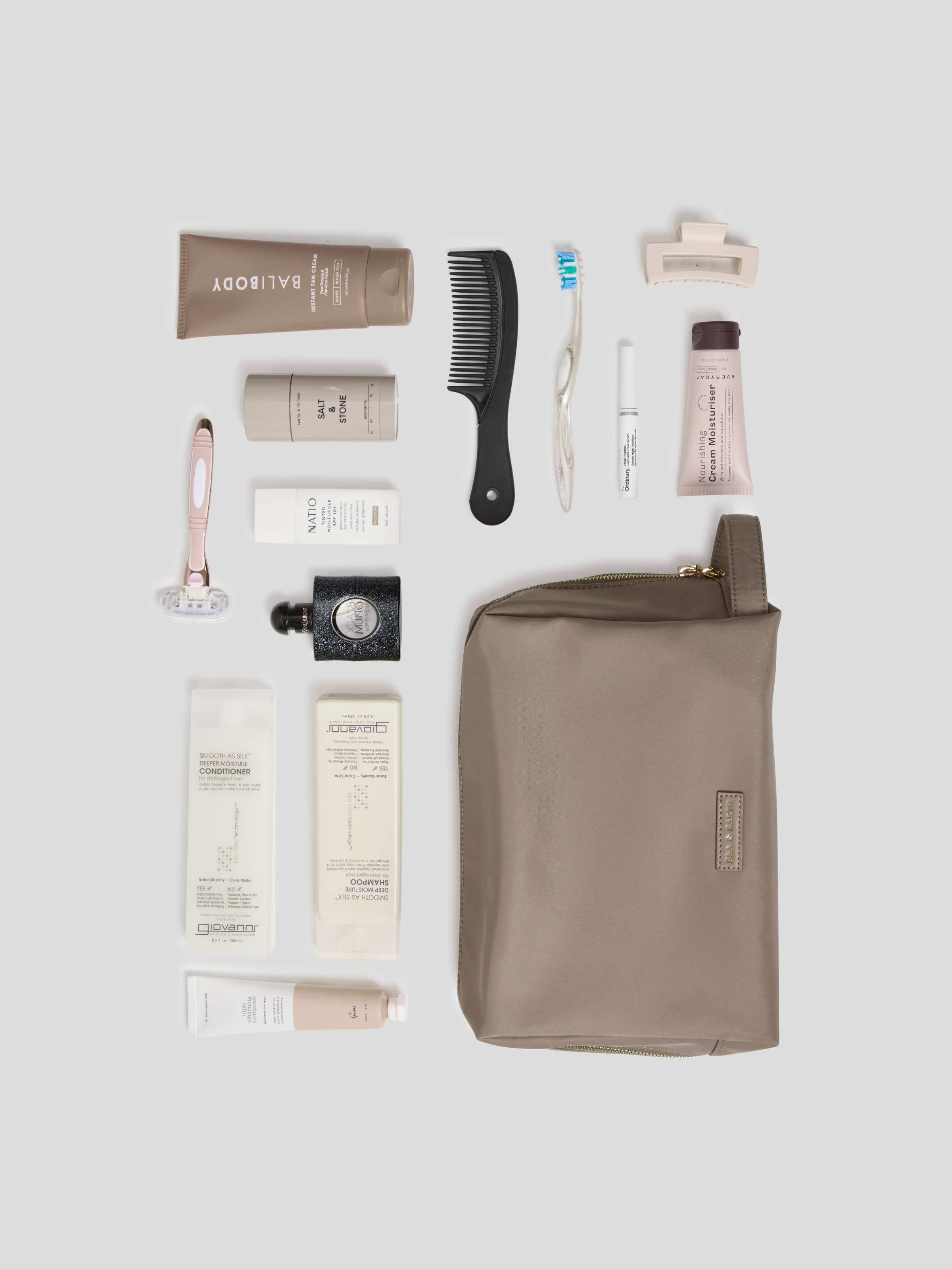 A flat-lay arrangement of toiletry and grooming items on a light gray background, including a taupe zip pouch, a black comb, a pink-handled razor, several white and beige tubes, and a small black bottle. The pouch is the largest item and appears to be the main product, with the other items styled as included accessories or companion products.