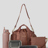 Rose  Lunch Bucket Bag set on a light gray studio background. The set includes a tan canvas duffel bag with a long shoulder strap, a matching tan pouch, a brown bottle, a dark gray lunch box, and a black lunch box with gold hardware